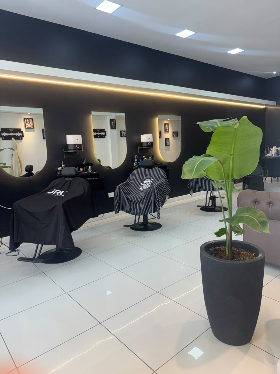 Interior de Flow BR Barber: tres sillones negros frente a espejos en forma de U con luz dorada, piso blanco y una planta en maceta negra.